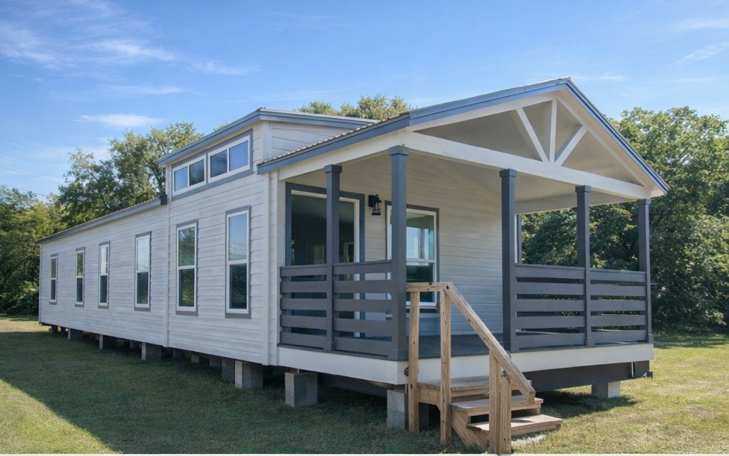 A modern tiny house with beige siding and dark trim sits on a grassy field. It features multiple windows and a small porch, exuding a cozy, inviting feel.