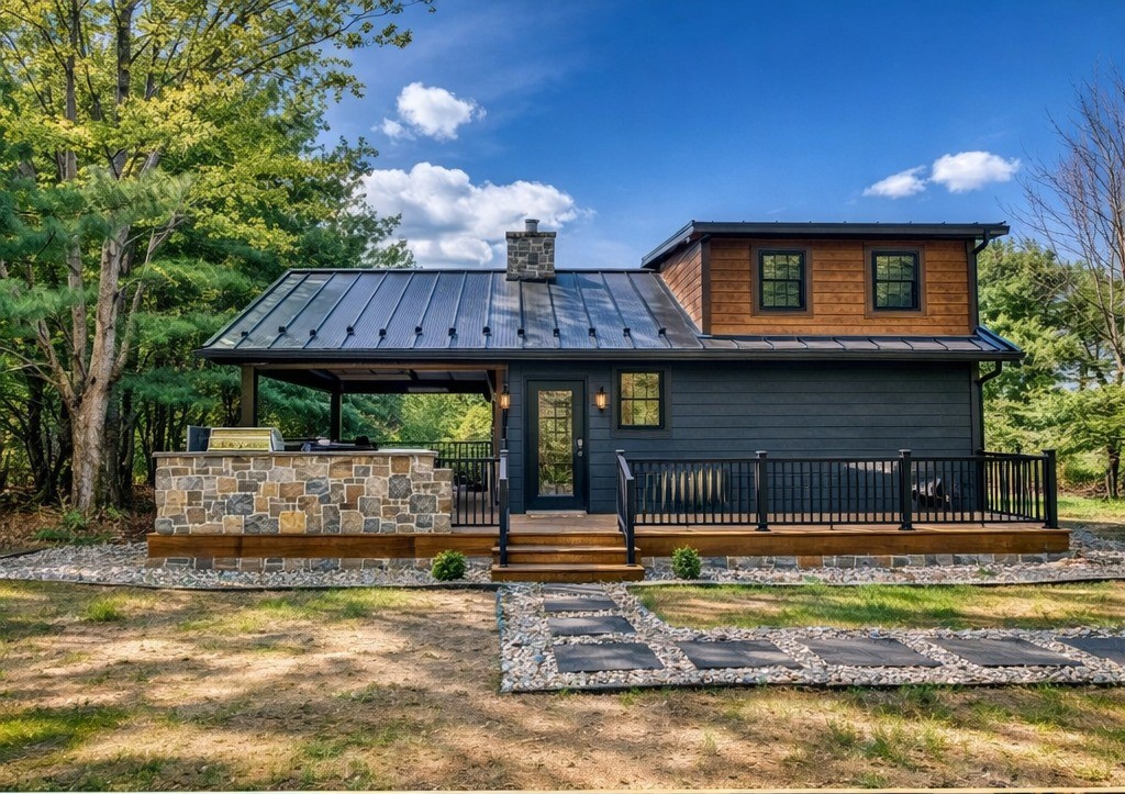 A modern cabin with dark wood siding and a metal roof sits among trees. It features a stone chimney, a front porch, and a stone pathway, under a clear blue sky.