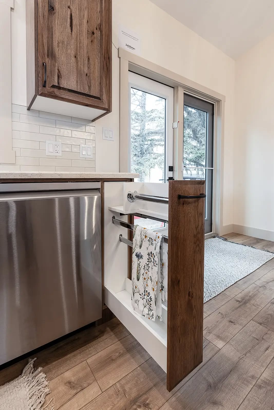 Kitchen with wood and stainless steel cabinets, open pull-out rack holding floral towels, beside glass doors leading to a snowy scene outside. Warm, modern feel.