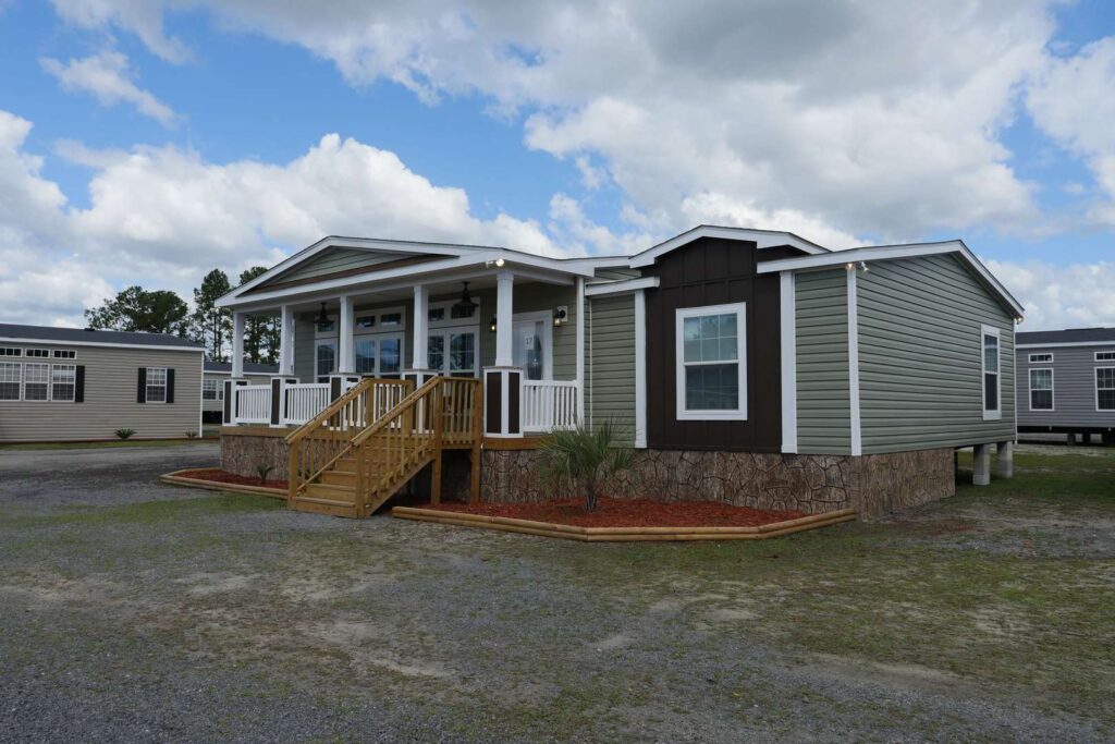 A single-story manufactured home features a sage green exterior with white trim. It has a covered front porch with wooden steps and a landscaped area. The sky is partly cloudy.