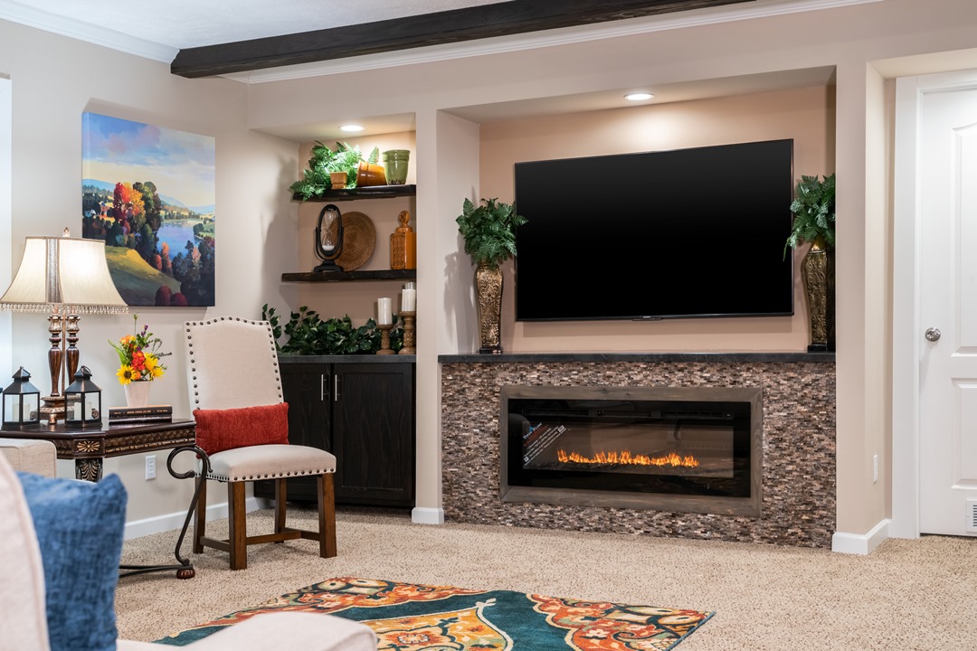 Cozy living room with a wall-mounted TV above a modern fireplace. Shelves with decor flank the TV. A colorful rug, lamp, and artwork add warmth.