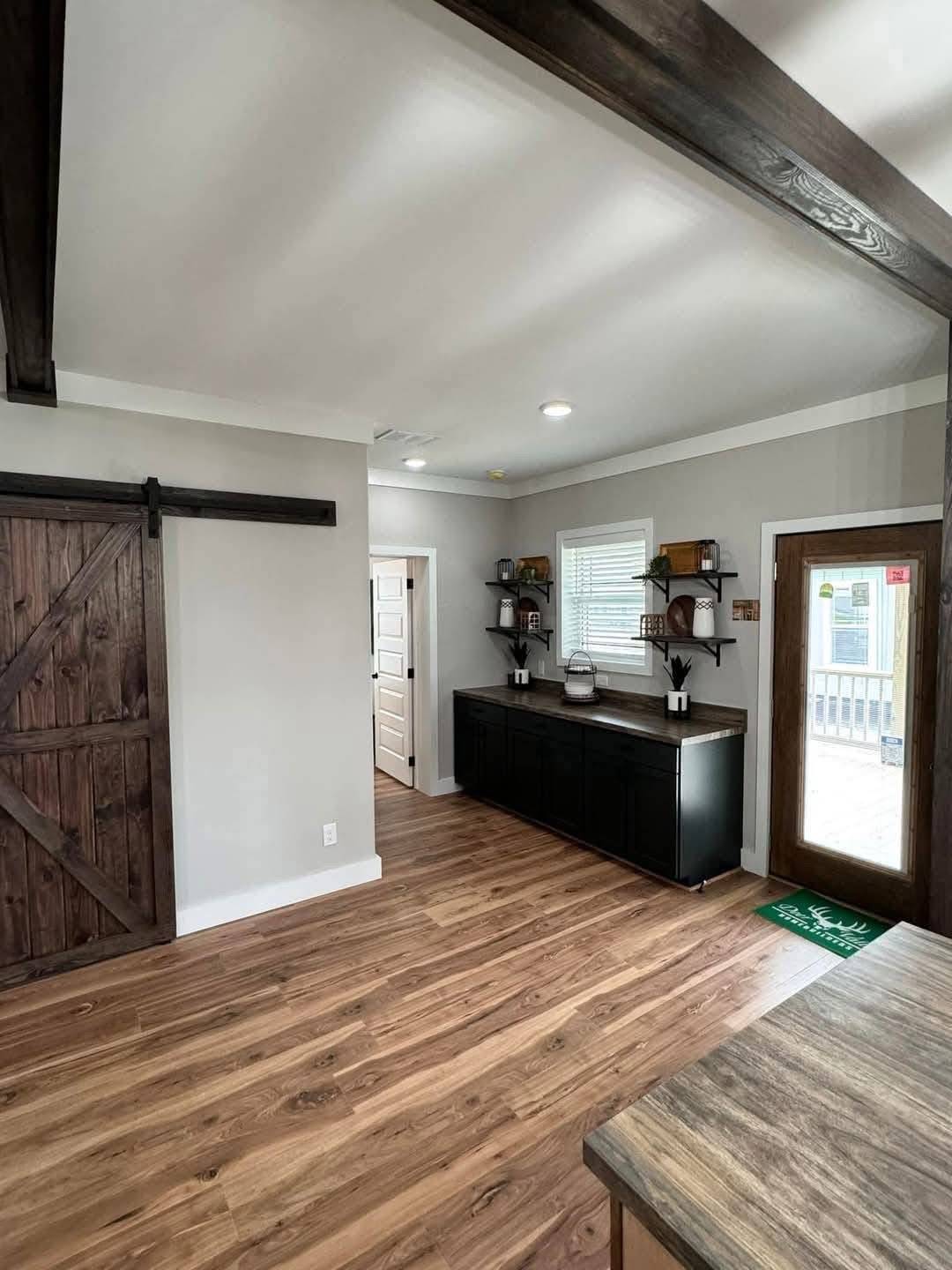 Modern interior with wood flooring and a rustic barn door on the left. Black counter with shelves and a doorway leading outside enhance a cozy atmosphere.