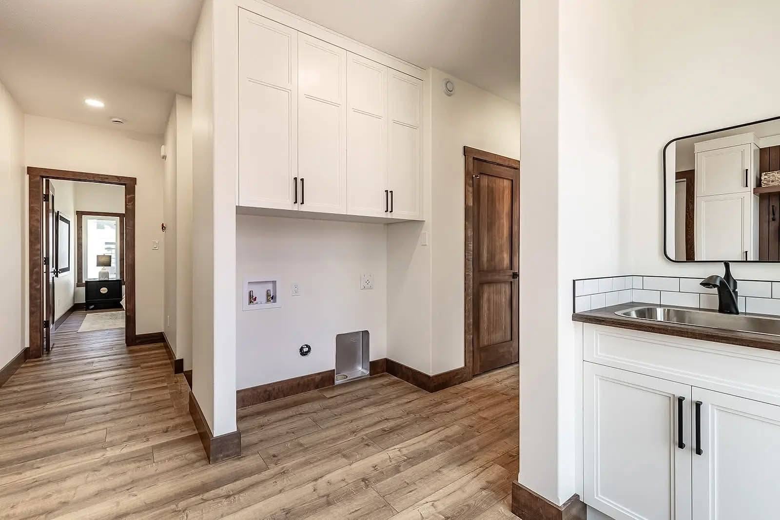 Laundry room with white cabinets and wood accents, featuring a space for appliances. A sink with a mirror is on the right, leading to a hallway on the left.