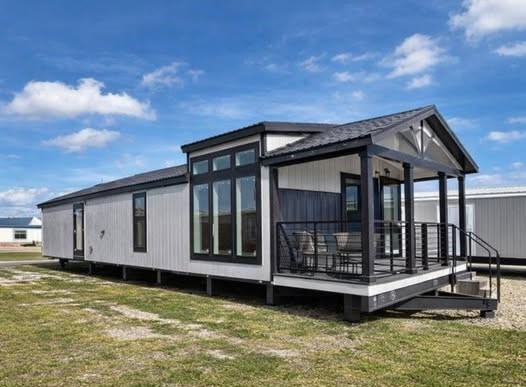 Modern mobile home under a blue sky, featuring large windows, a small covered porch with chairs, and a black railing, on a grassy lot.