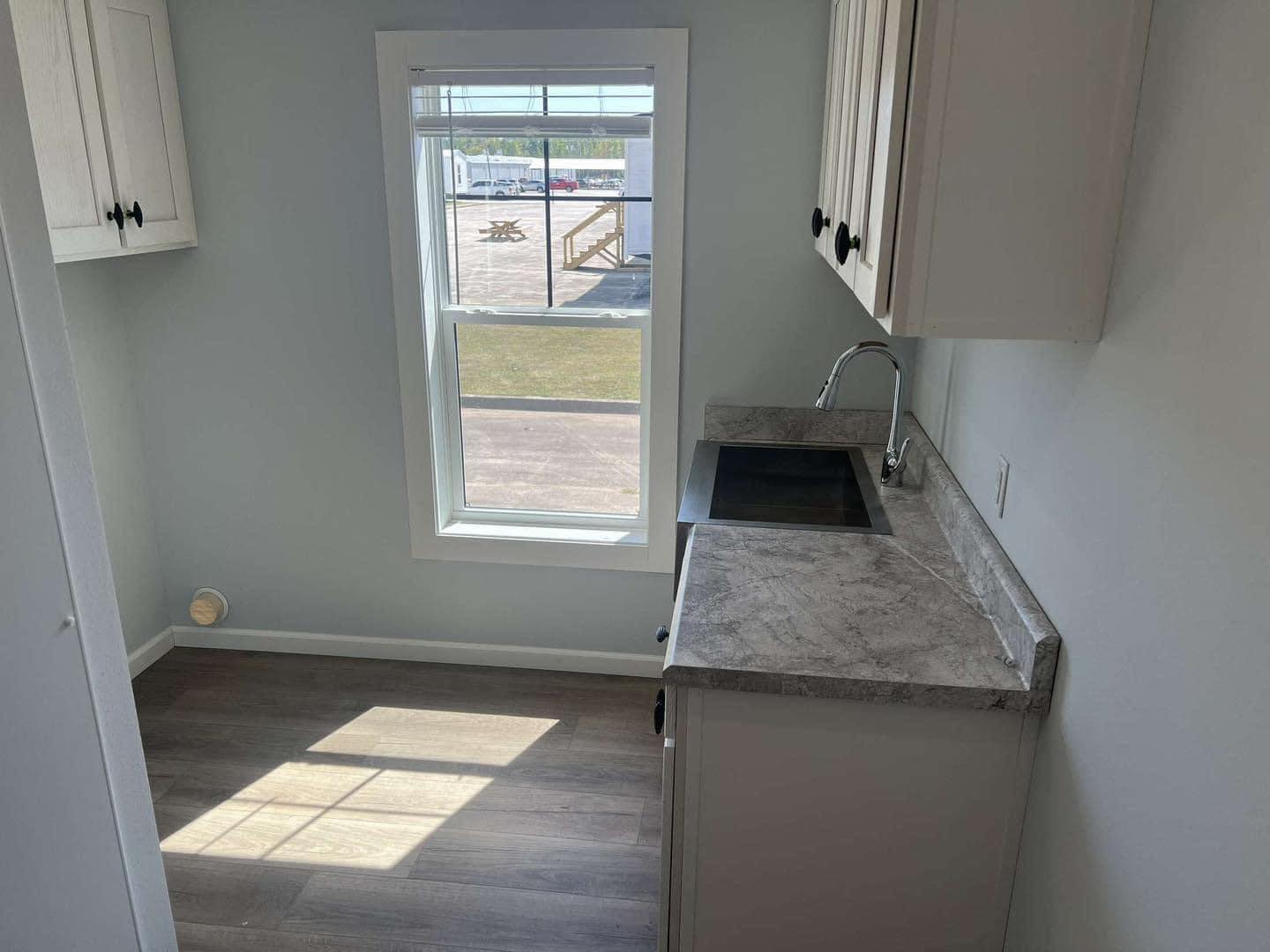 Compact kitchen with light gray walls and wooden floor. Sunlight streams through a window above the counter, illuminating a sink with a chrome faucet.
