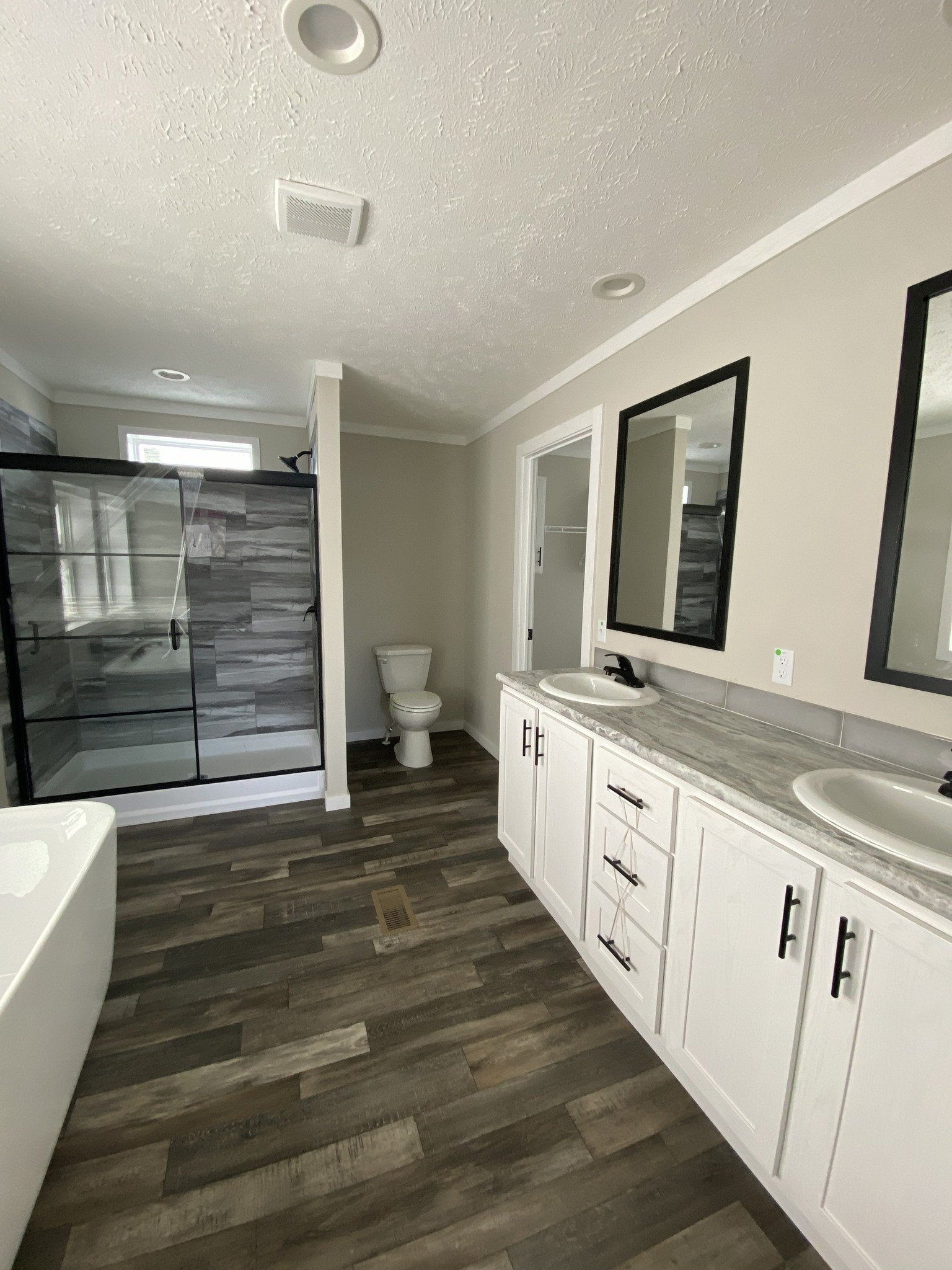 Spacious modern bathroom with dual sinks on a gray countertop, two black-framed mirrors, a wooden floor, a glass shower, and a toilet in the back.