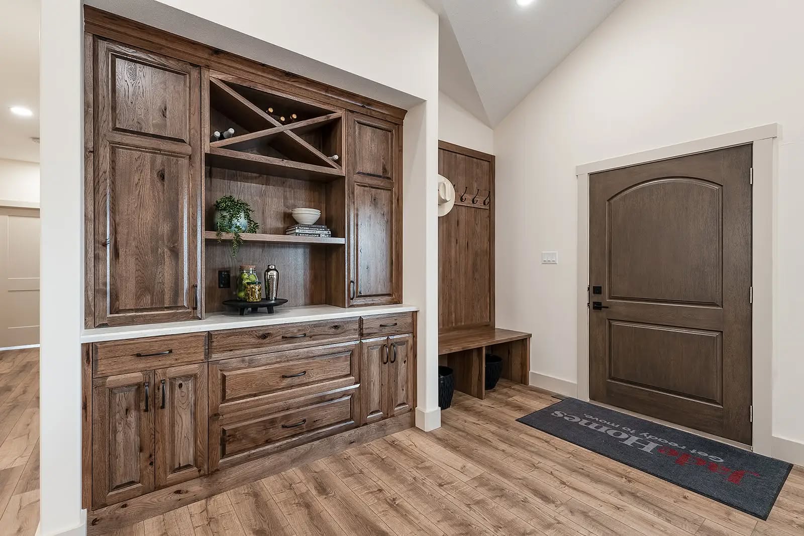 Spacious entryway with rustic wooden cabinetry, a bench with coat hooks, and a closed wooden door. Light wood flooring and a welcoming rug.