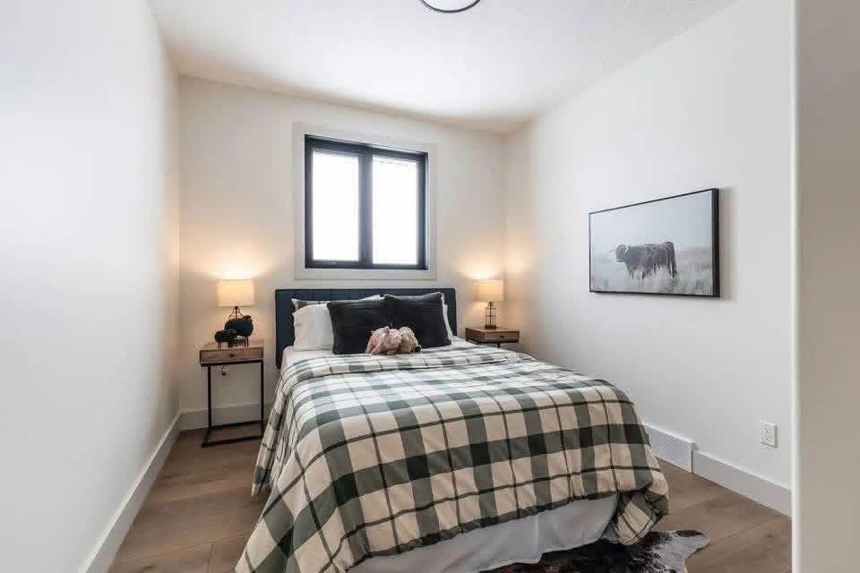 Cozy bedroom with a checkered bedspread, flanked by two nightstands with lamps. A window above the bed and a cow-themed artwork add a rustic touch.