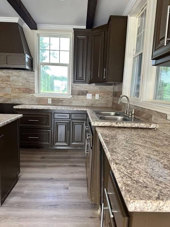 Modern kitchen with dark brown cabinets, marble countertops, and wood flooring. Large windows provide natural light, creating a warm, inviting atmosphere.