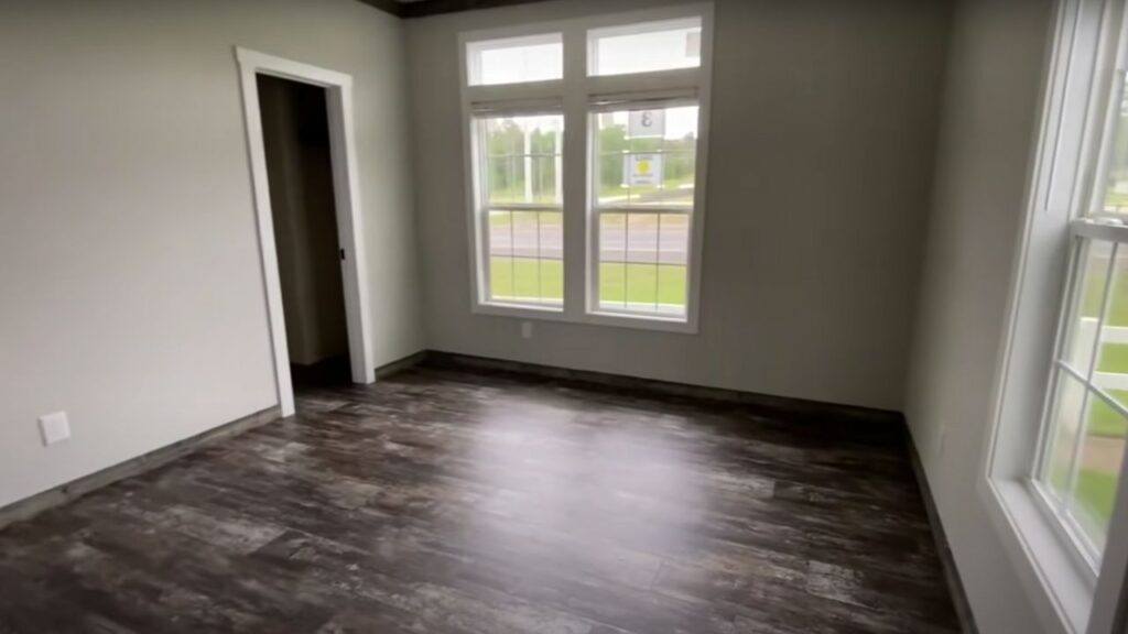 An empty room with dark wood flooring and white walls. It features two large windows letting in natural light, and an open doorway on the left.
