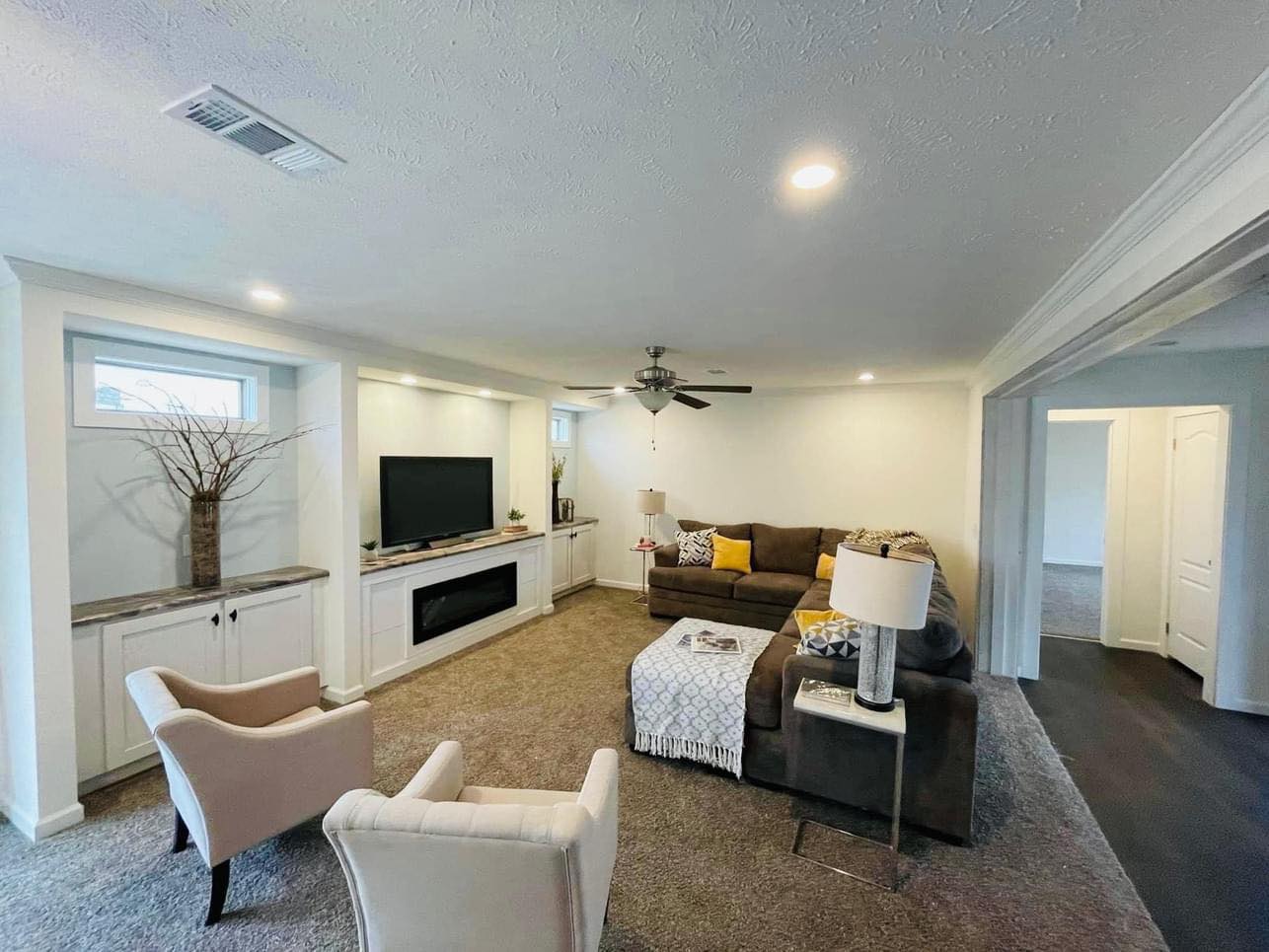 Cozy living room with a sectional sofa, decorative pillows, and a plaid throw. Two armchairs face a TV on a built-in shelf. Soft lighting and neutral tones.