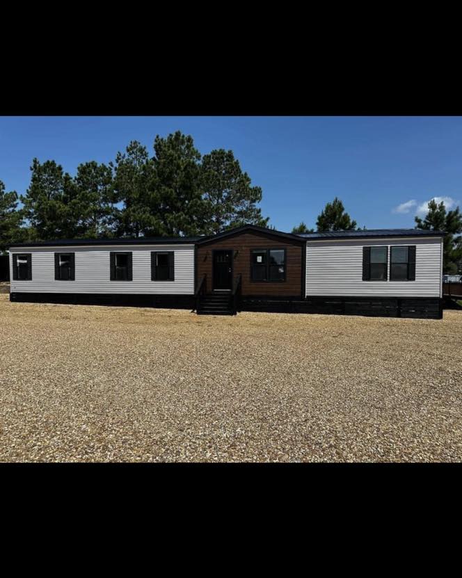 A single-story mobile home with a dark roof and a central brown panel entrance is set on a gravel lot. Tall green trees stand under a blue sky.