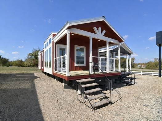 A red tiny house with white trim and a modern porch sits on gravel under a clear blue sky. The bright setting gives a welcoming, cozy feel.
