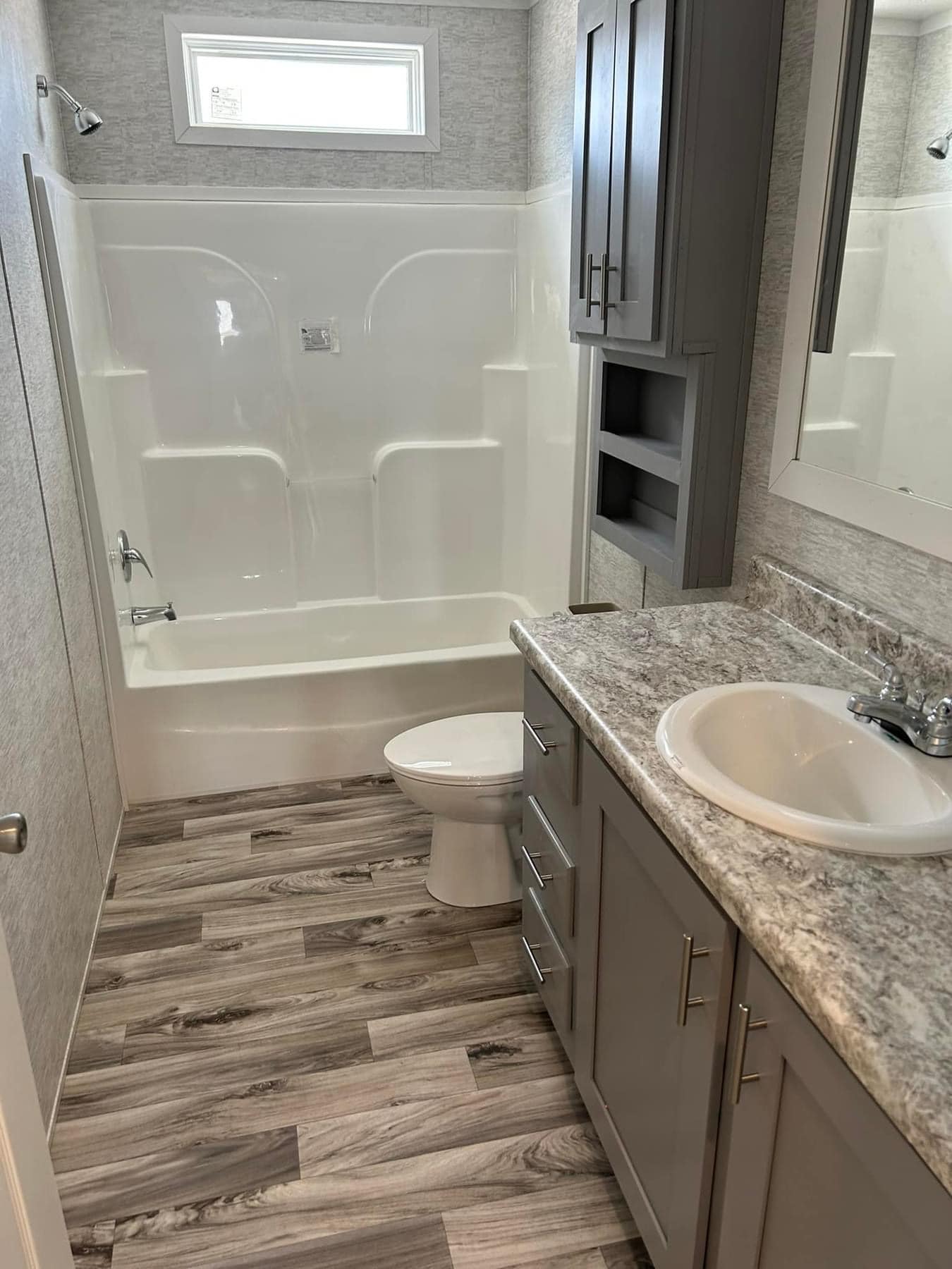Modern bathroom with gray cabinets, marble-patterned countertop, and wood-like flooring. A white bathtub is next to the toilet, enhancing the clean look.
