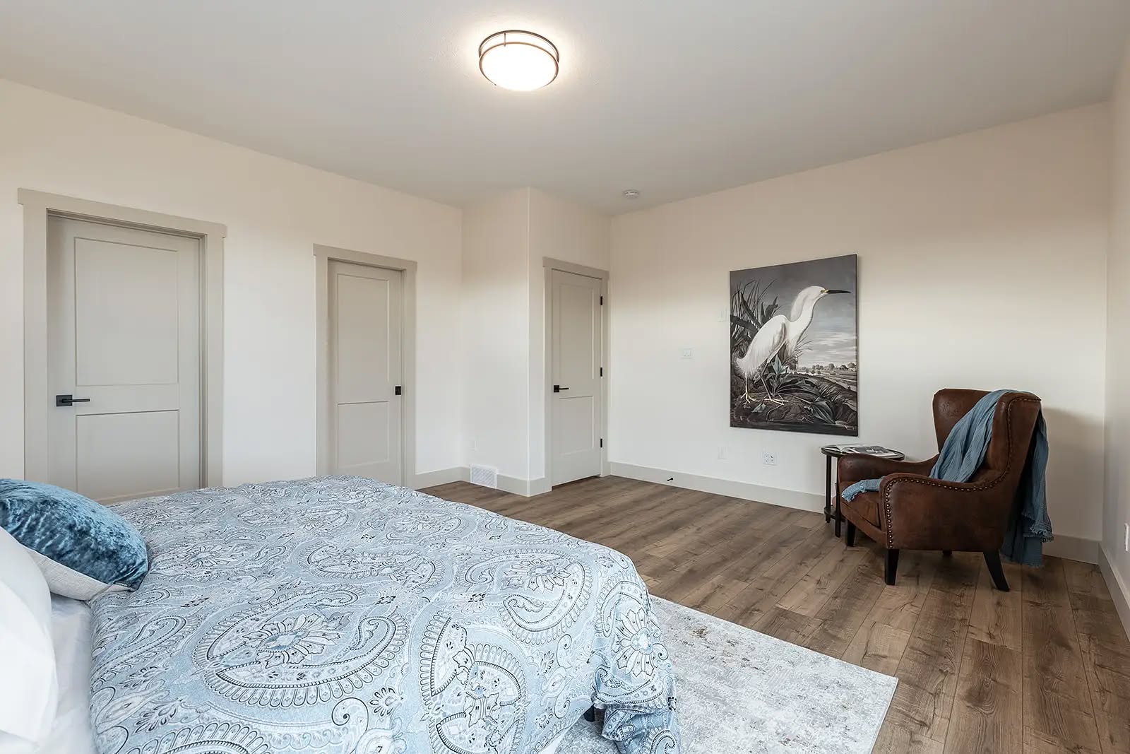 A serene bedroom with a light blue paisley-patterned bedspread, light wood flooring, a brown leather chair with a blue throw, and a bird painting on the wall.