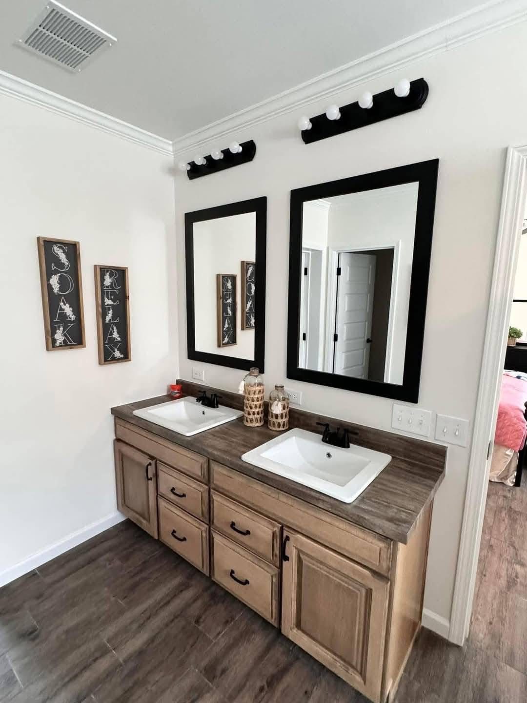 Wooden vanity with two white sinks under black-framed mirrors in a bathroom. Decor includes "Soak" and "Relax" signs and jar accents, creating a serene tone.