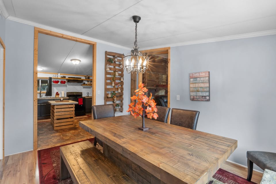 Dining room with a rustic wooden table, orange flower centerpiece, and a vintage chandelier. Kitchen visible through doorway; cozy and inviting atmosphere.