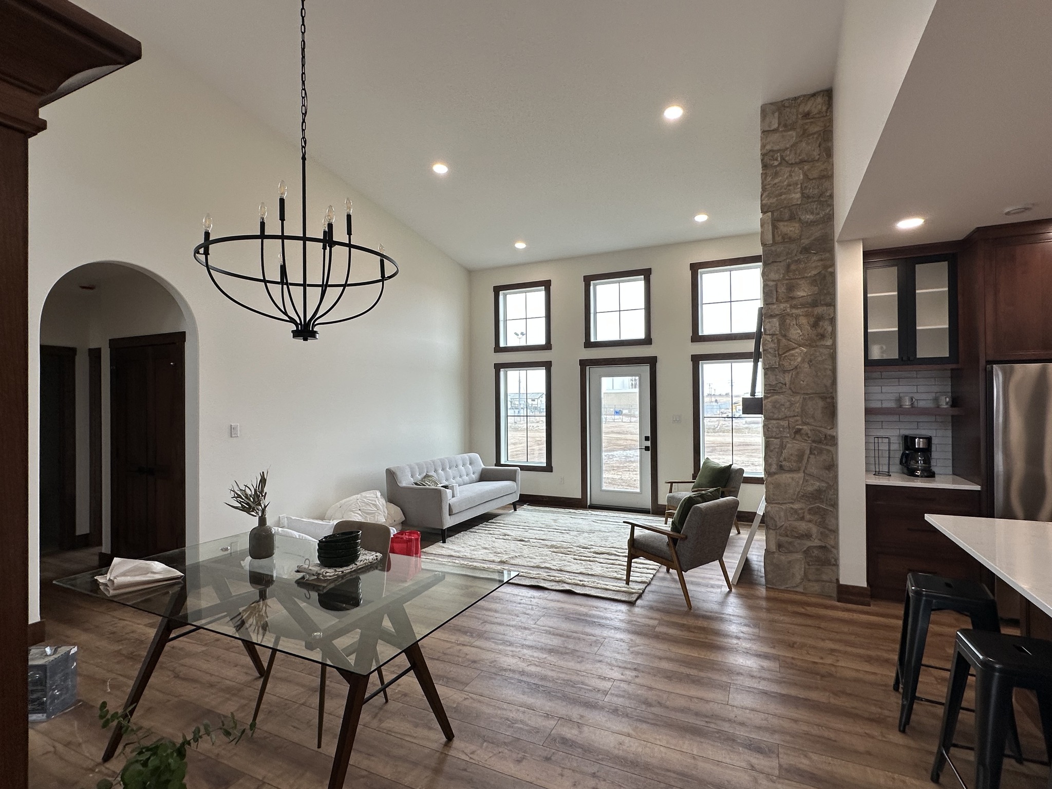 A modern living room with large windows, a light gray sofa, and an armchair. Stone column detail, glass dining table, dark wood, and airy atmosphere.