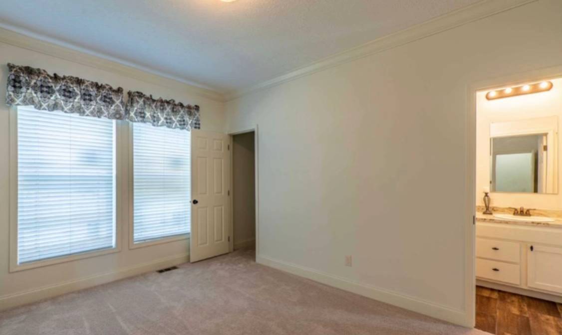 A neutral-toned empty room with carpet, two large windows, and a patterned valance. An open door leads to a bright bathroom with a lit mirror. Cozy and inviting atmosphere.