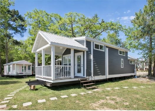 A modern tiny house with a dark gray exterior and white trim sits surrounded by trees. It features a small porch and stepping stones on a grassy lawn.