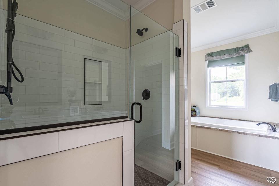 Spacious bathroom featuring a glass-enclosed shower with white tile, adjacent to a sunlit bathtub under a window. Modern design with a calm atmosphere.