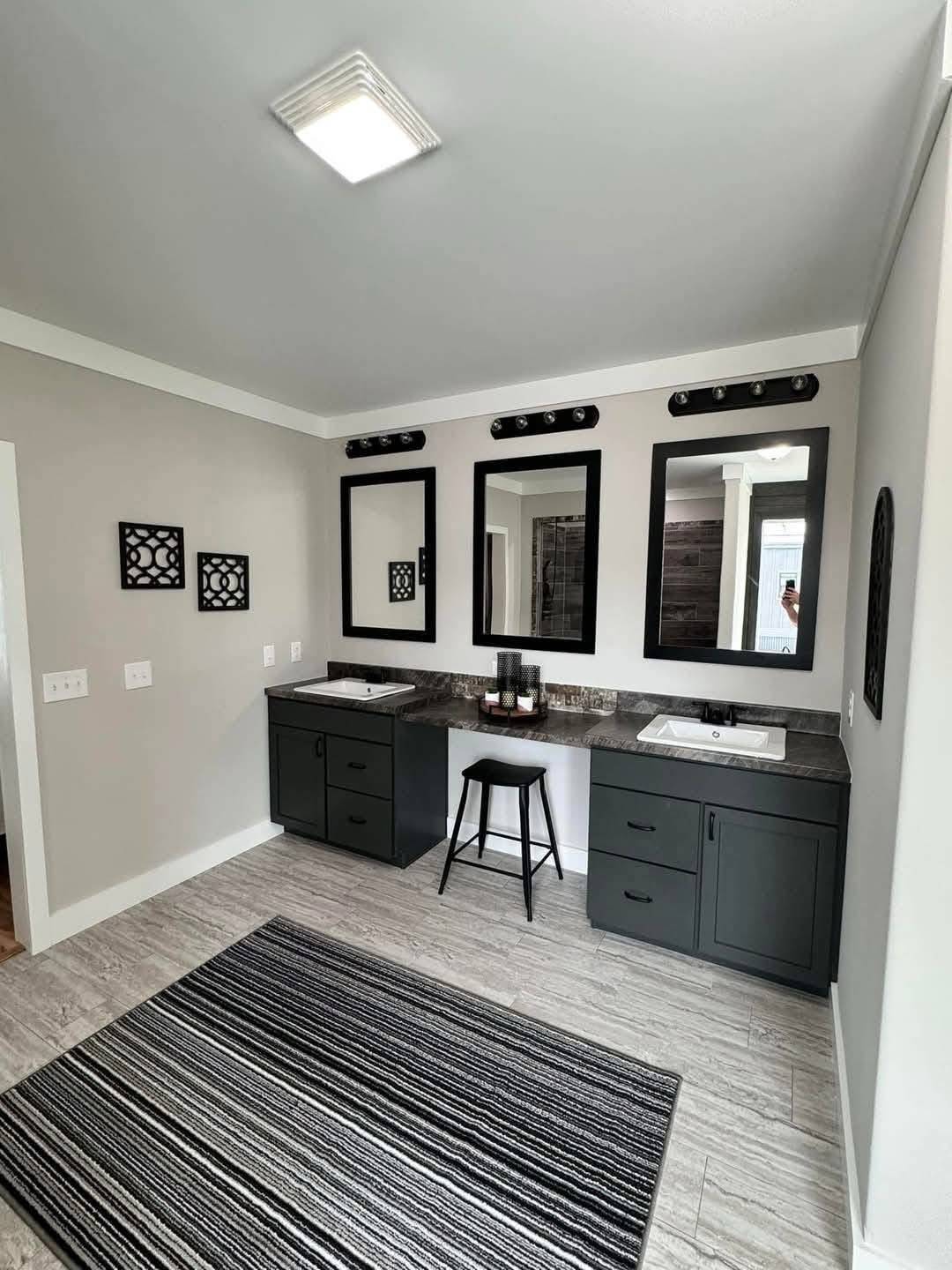 Modern bathroom with dual sinks and dark green cabinets. Three large mirrors above a charcoal countertop. A striped rug on light tile flooring.