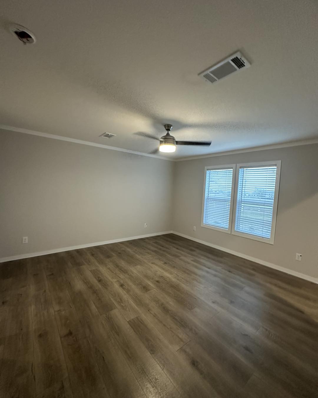 Spacious empty room with light gray walls and brown wooden floor. Ceiling fan with light hangs overhead. Two windows with blinds bring in soft light.