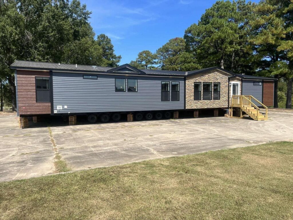 A large manufactured home on wheels sits on a concrete lot with grass in the foreground. It has blue siding and a stone accent around the door, with trees in the background.