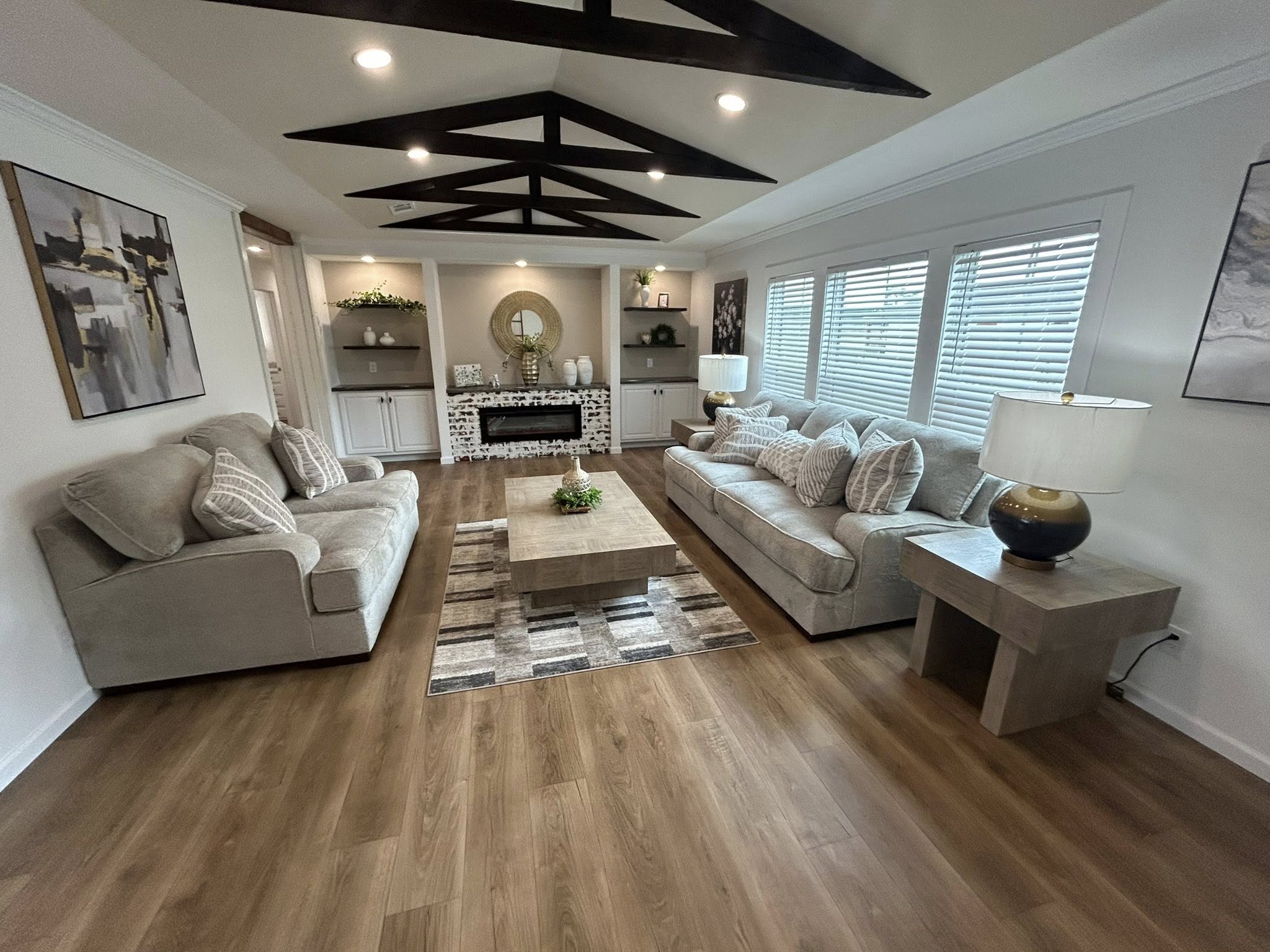 Stylish living room with wood floor, beige sofas, patterned cushions, central coffee table, art on walls, modern lighting, and decorative shelves.