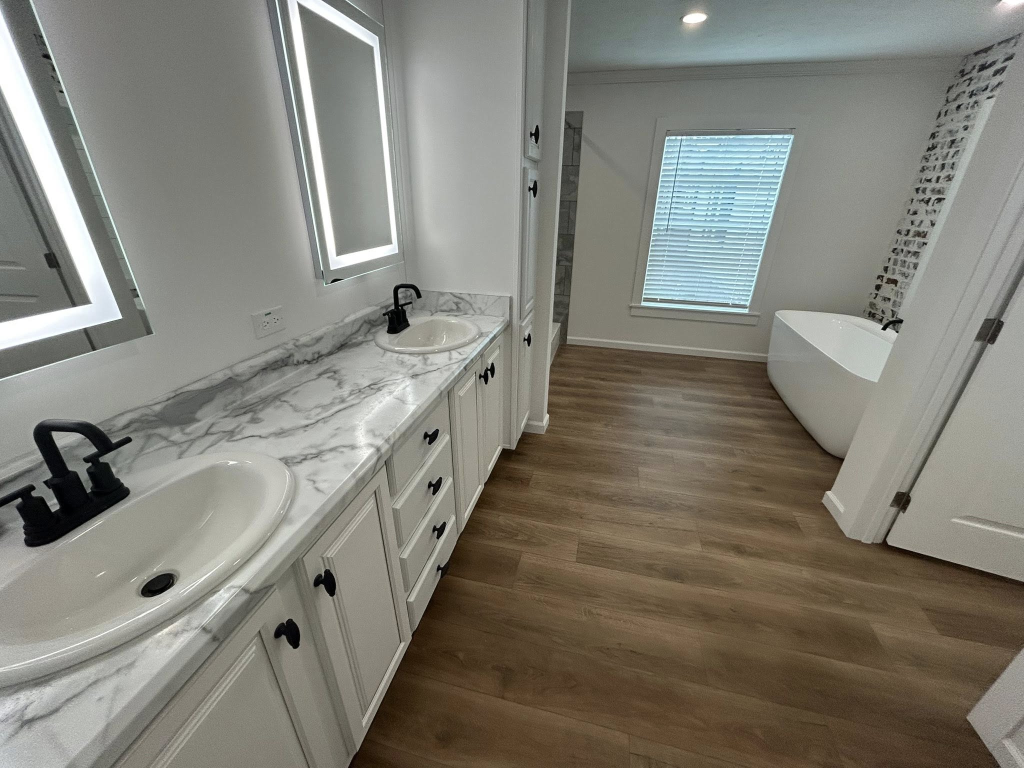 Stylish bathroom with marble countertops, two sinks, and black fixtures. Accented by illuminated mirrors, wood flooring, and a freestanding tub. Cozy elegance.