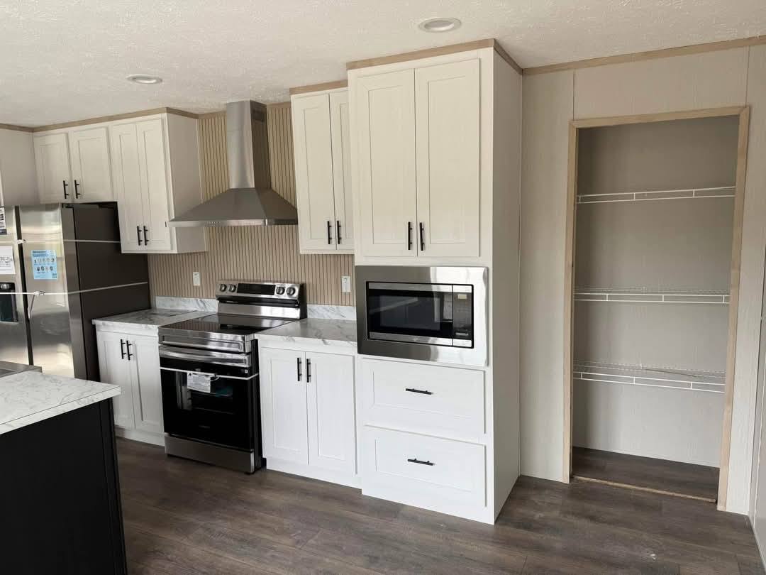 Modern kitchen with white cabinets, stainless steel appliances including a fridge, oven, and microwave. Open pantry with wire shelves. Warm, minimal decor.