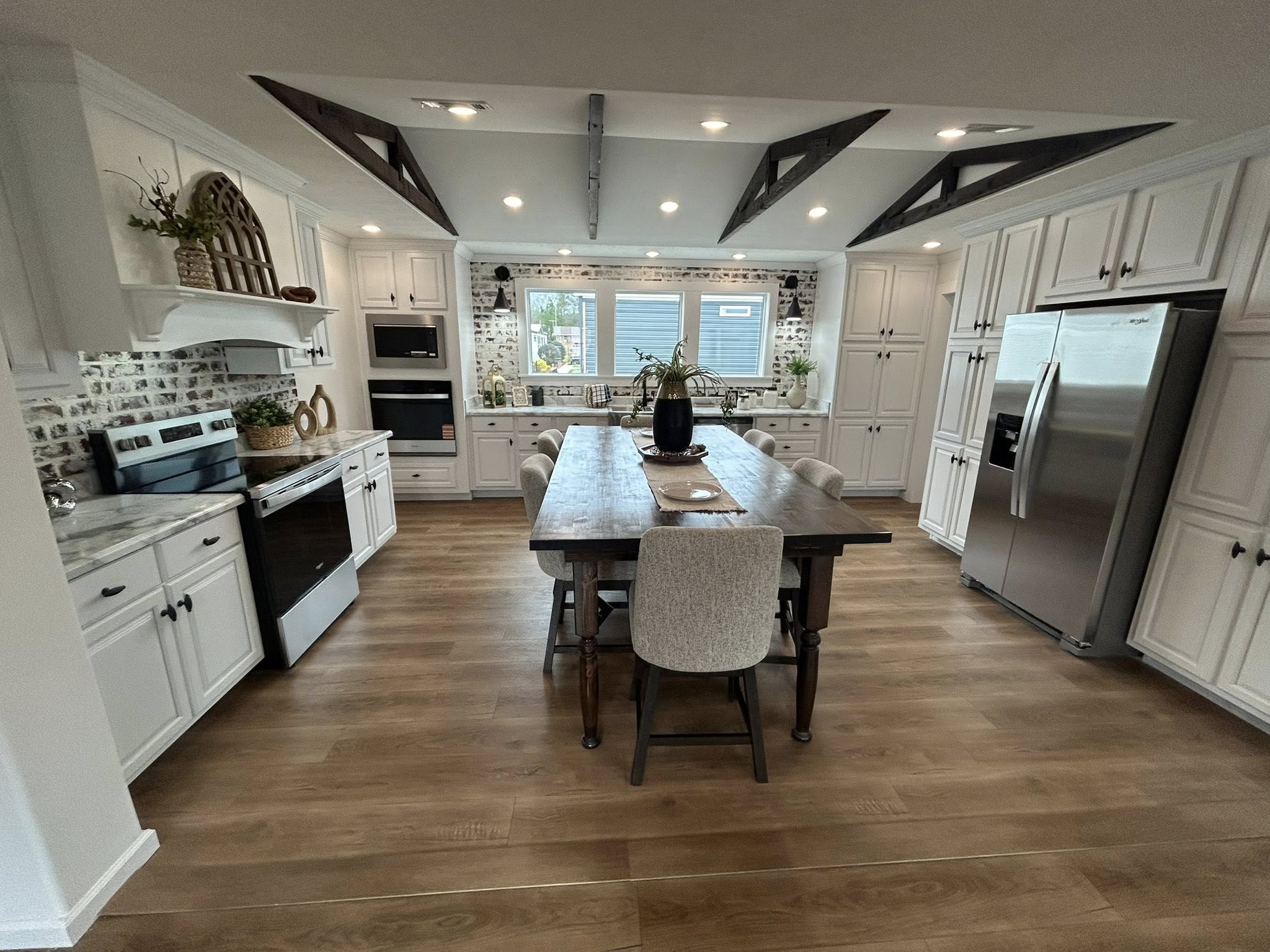 Spacious kitchen with white cabinets, brick backsplash, and wood floor. Central wooden island with chairs, stainless steel fridge, and decorative beams.
