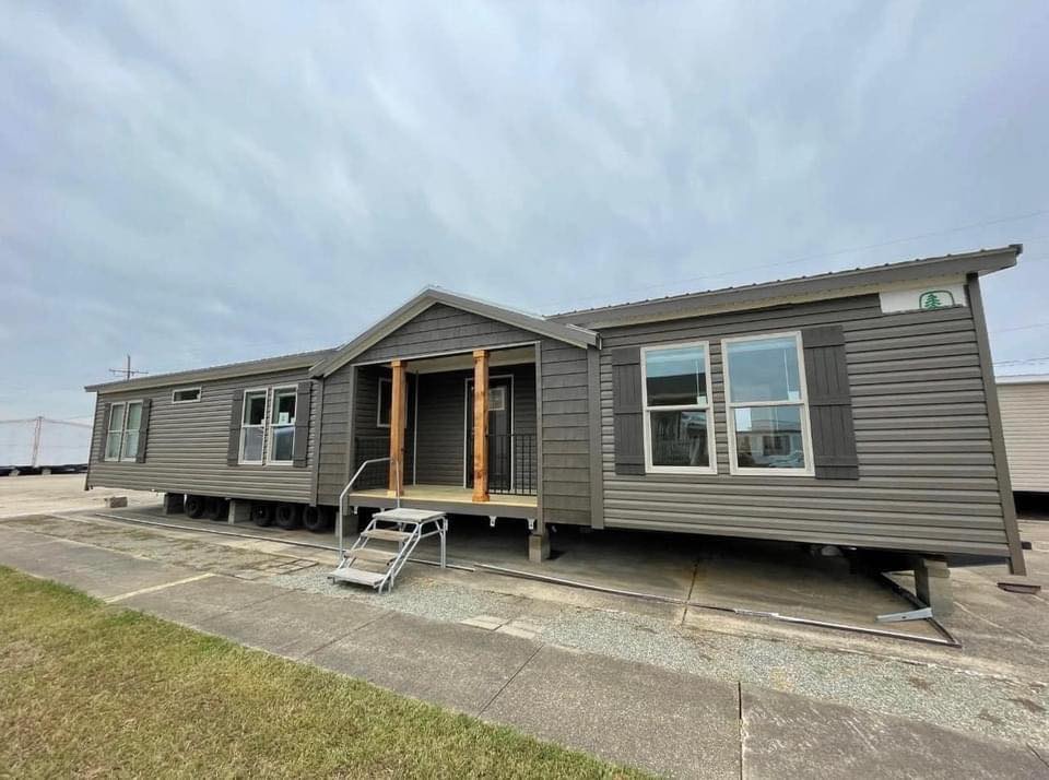 A large gray mobile home on a lot, featuring a small porch with wooden columns and metal steps, under an overcast sky.
