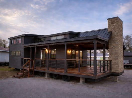 Modern tiny house with dark wood exterior and large stone chimney on wheels. Wide porch with railings, surrounded by twilight sky. Cozy ambiance.