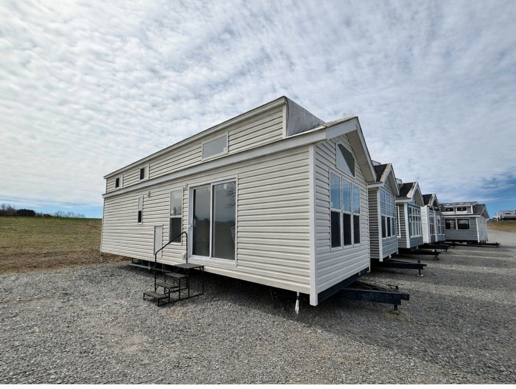 Row of uniform white tiny homes with large windows on gravel lot under a cloudy sky, conveying a sense of new development and simplicity.