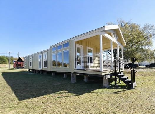 Modern beige modular home on raised foundation with large windows and a small porch. Set in a grassy area under a clear blue sky, evoking a peaceful atmosphere.