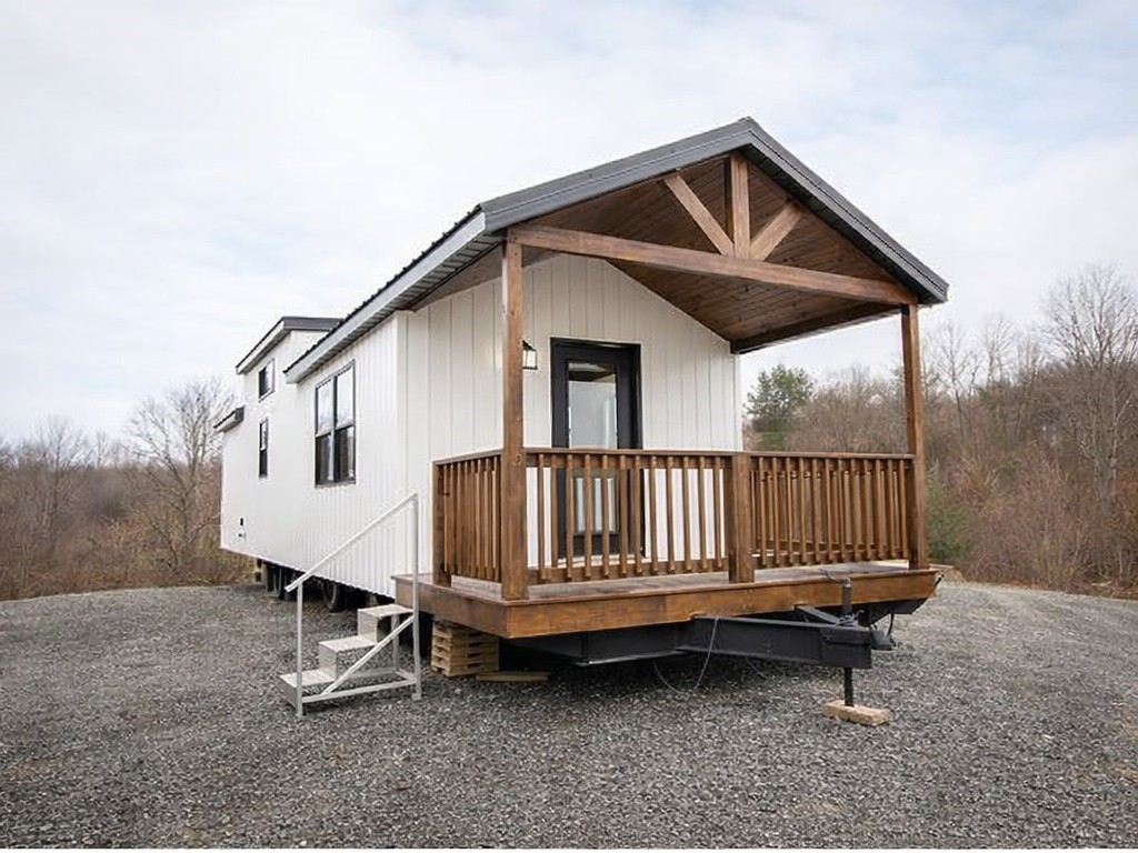 A modern tiny house with white siding and a wooden porch stands on a gravel lot. It is surrounded by leafless trees and under a cloudy sky, conveying a serene, rustic vibe.