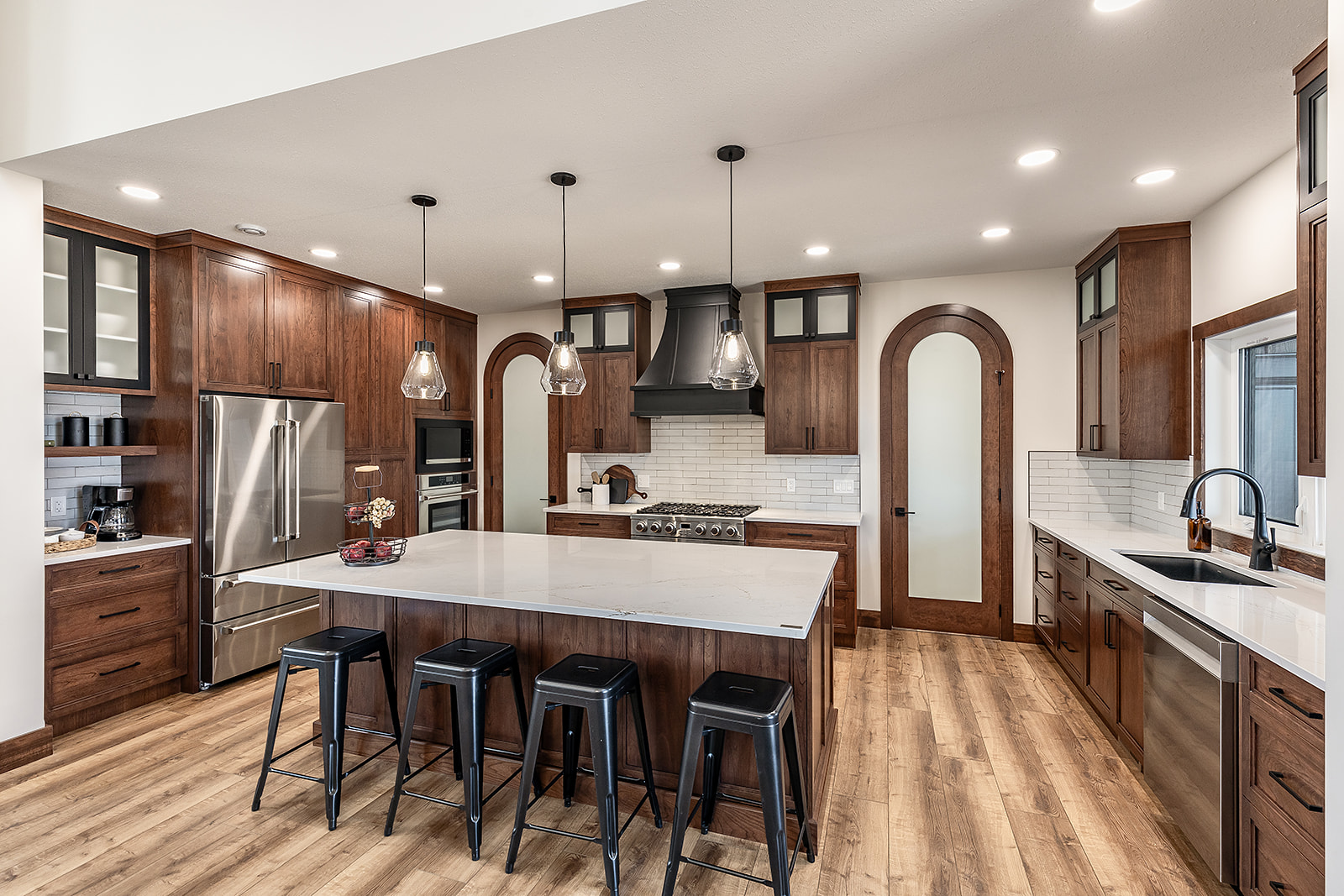 Modern kitchen with warm wood cabinets, large island with white countertop, and black barstools. Stainless steel appliances and pendant lighting.
