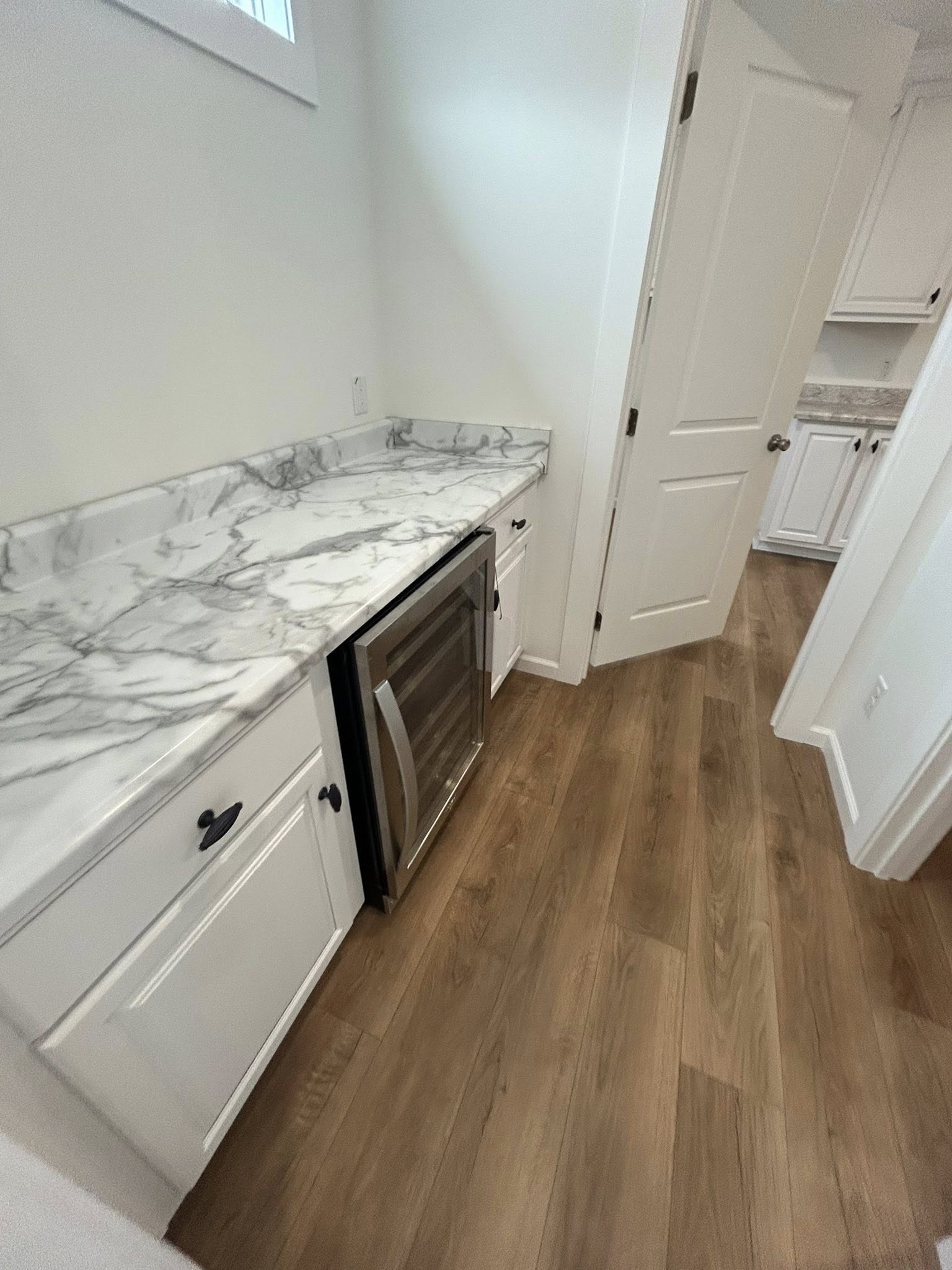 A laundry room featuring white cabinets with black handles, a marble-patterned countertop, a stainless steel wine fridge, and wood flooring.