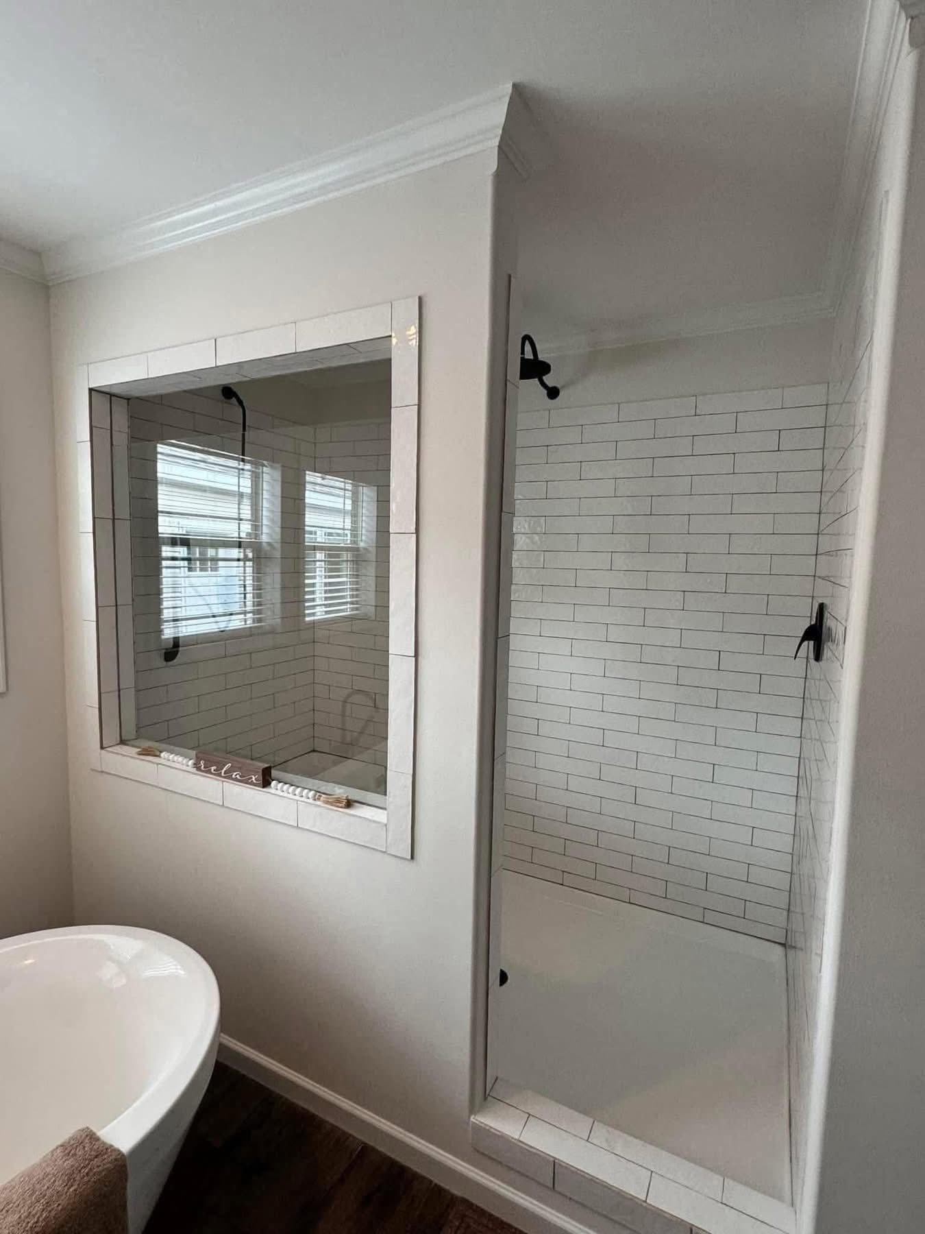 A modern bathroom features a white tiled shower with black fixtures, a large mirror reflecting windows, a freestanding bathtub, and a warm wood floor.