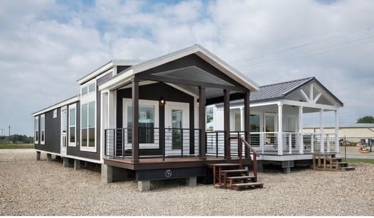 Two modern tiny homes with large windows and porches sit on a gravel lot under a cloudy sky. The houses are elevated with wooden steps leading up.