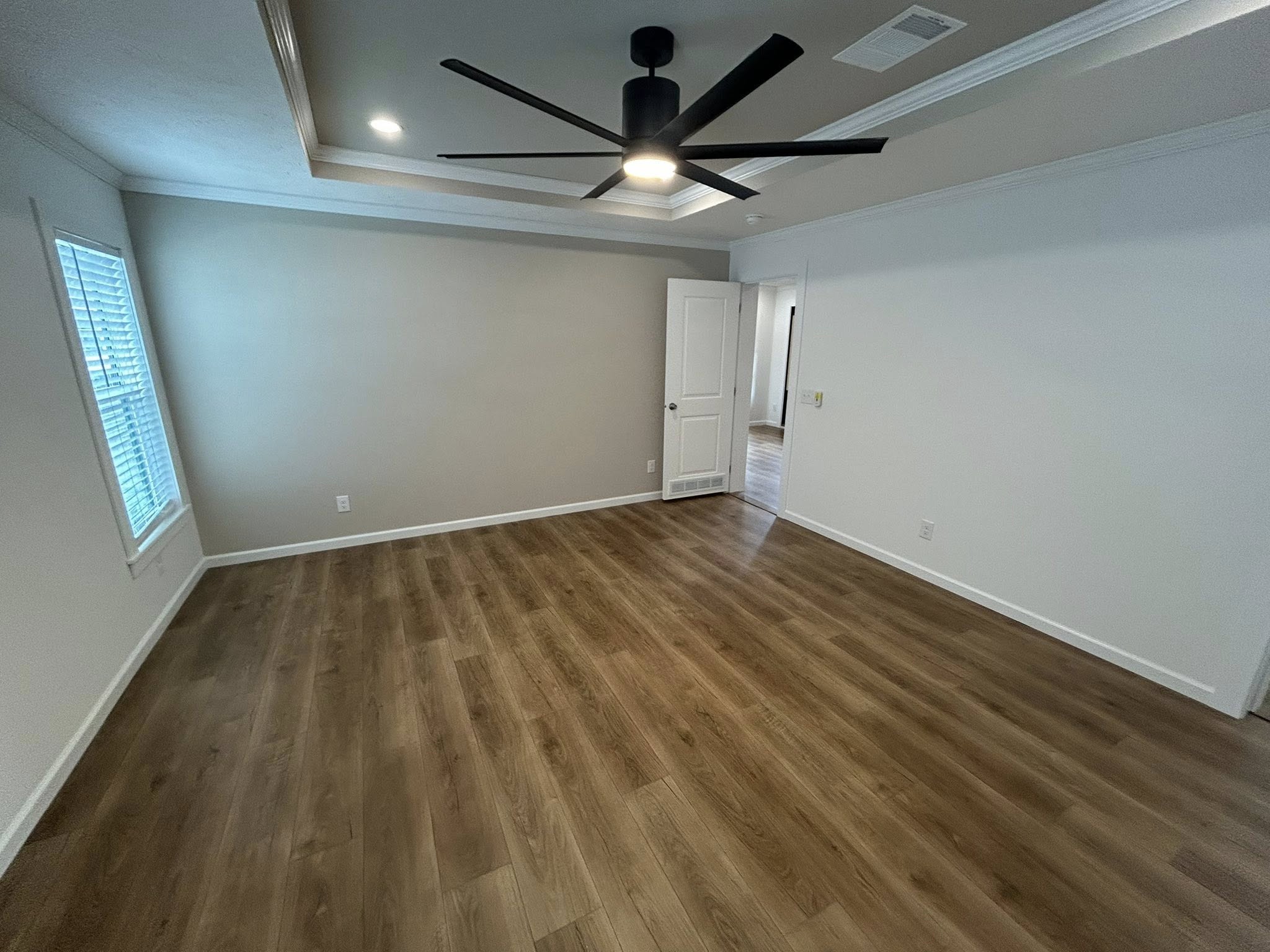 Spacious empty room with wood flooring, beige walls, a white ceiling, and a large black ceiling fan. Natural light enters through a window on the left.