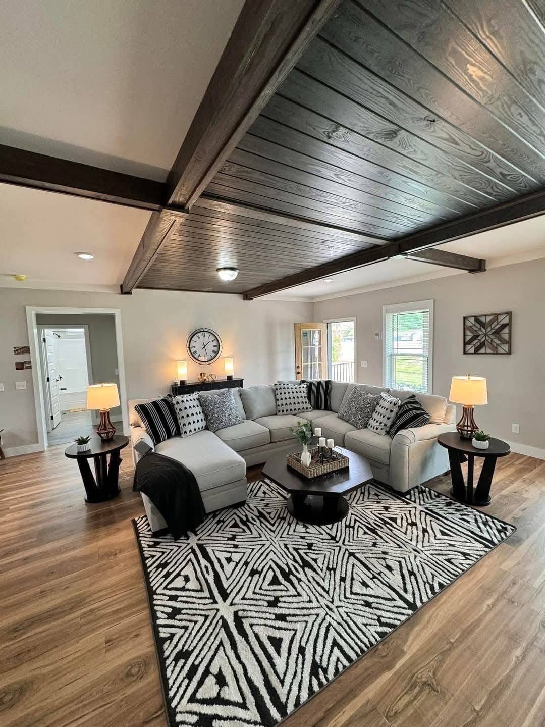 A cozy living room with a gray sectional sofa adorned with patterned cushions. A geometric rug sits on wooden flooring under a wooden beam ceiling. Two lamps on side tables emit a warm glow, enhancing the welcoming atmosphere. A wall clock and window add to the modern decor.