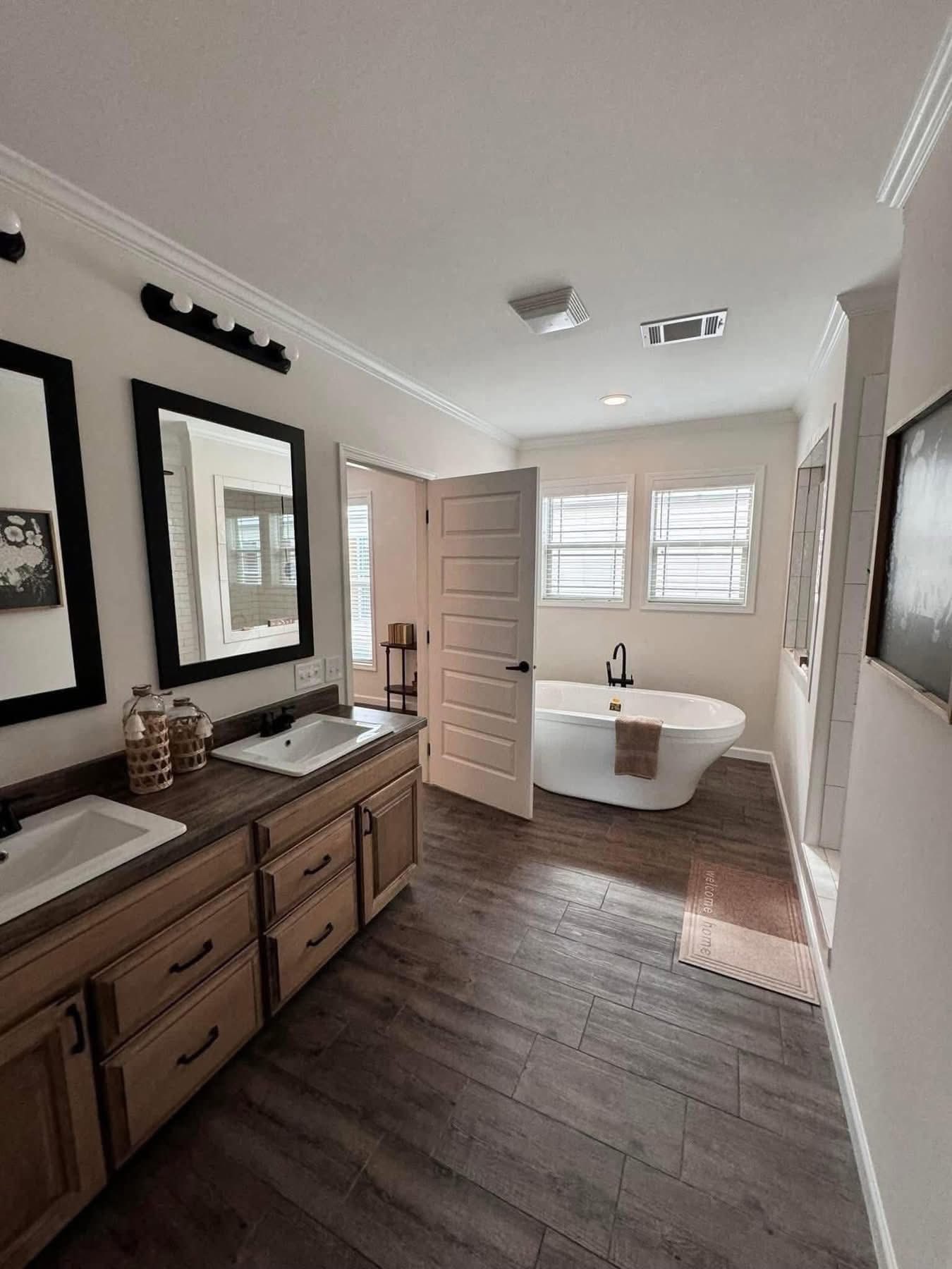 Modern bathroom with wooden cabinets and dual sinks on the left. A freestanding tub with a towel is on the right, under three windows. Calm ambiance.