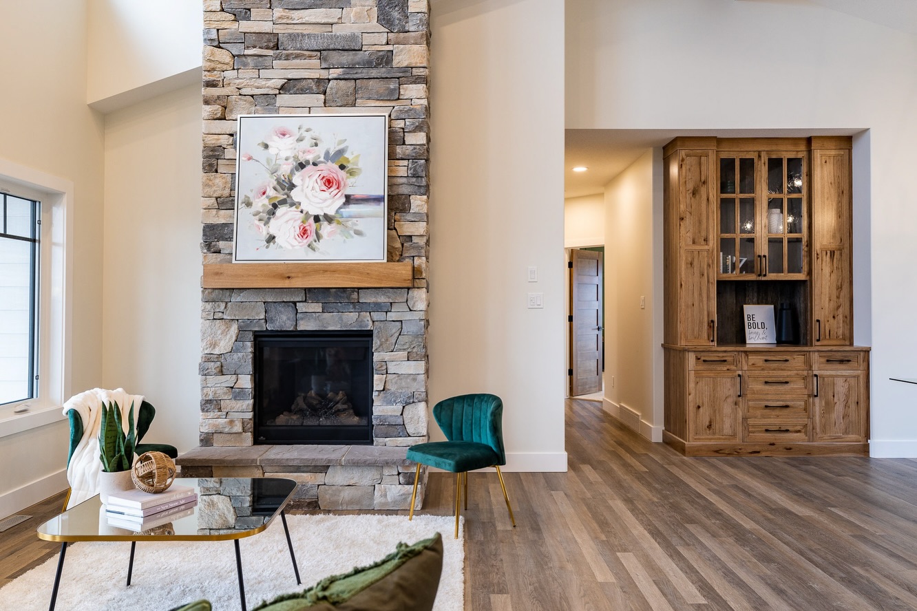 Cozy living room with a stone fireplace, floral art, and elegant decor. Green velvet chairs and wood accents create a warm, welcoming ambiance.