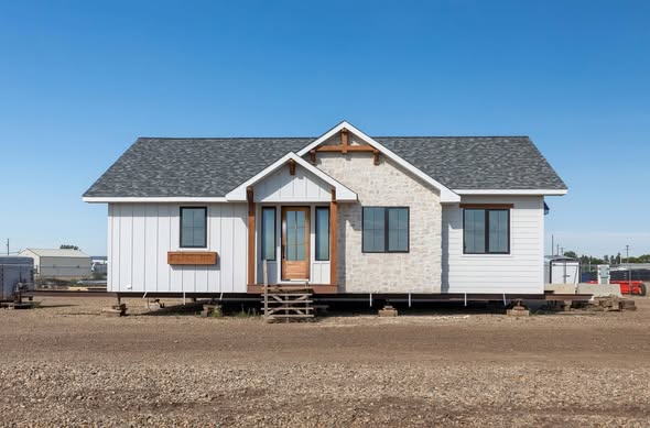 A charming, small white house with a gray shingle roof sits on a gravel lot in a rural area. Its design is simple and neat, giving a cozy feel.