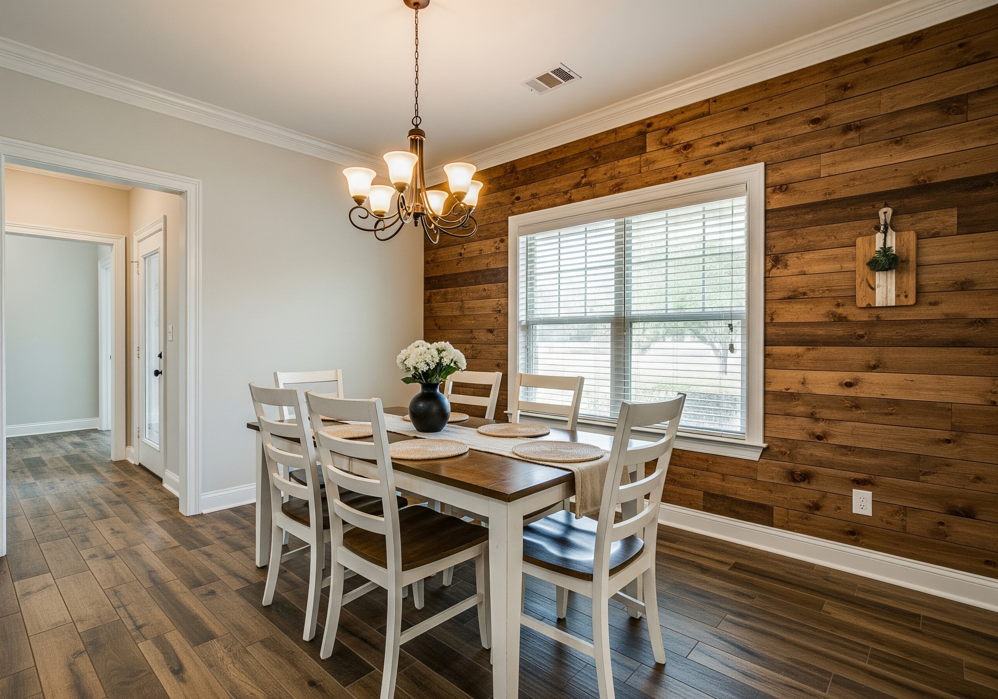 A cozy dining room features a wooden table with six white chairs, a black vase with flowers, a wooden accent wall, and a warm chandelier above.