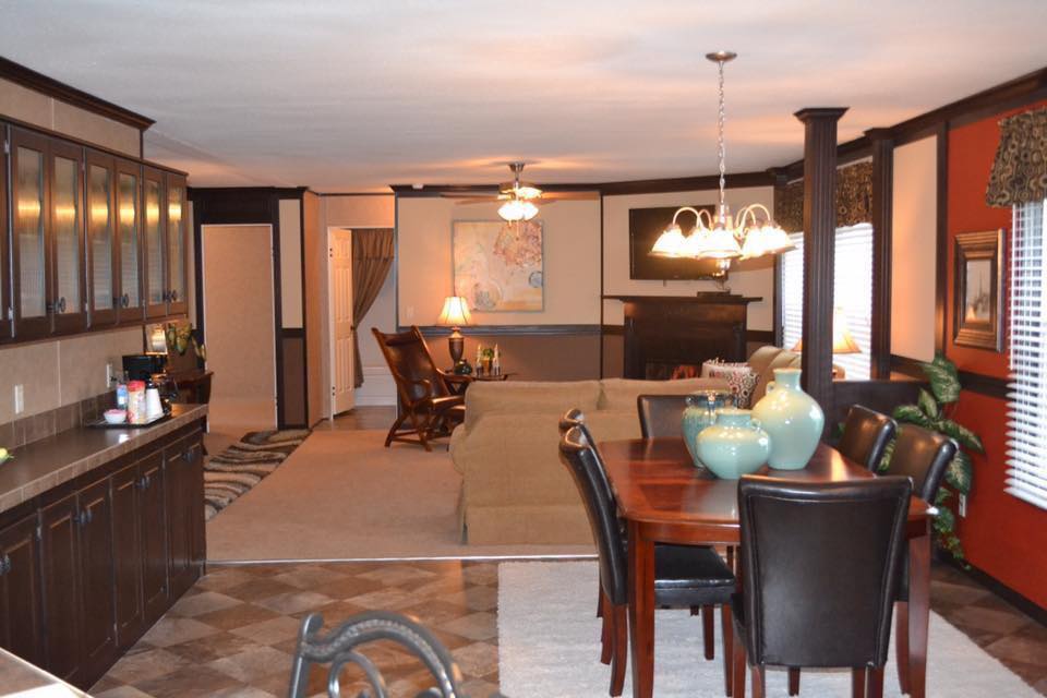 Warmly lit living and dining area with dark wood accents. Features a large table, leather chairs, and plush sofas. Decorative vases and framed art enhance coziness.