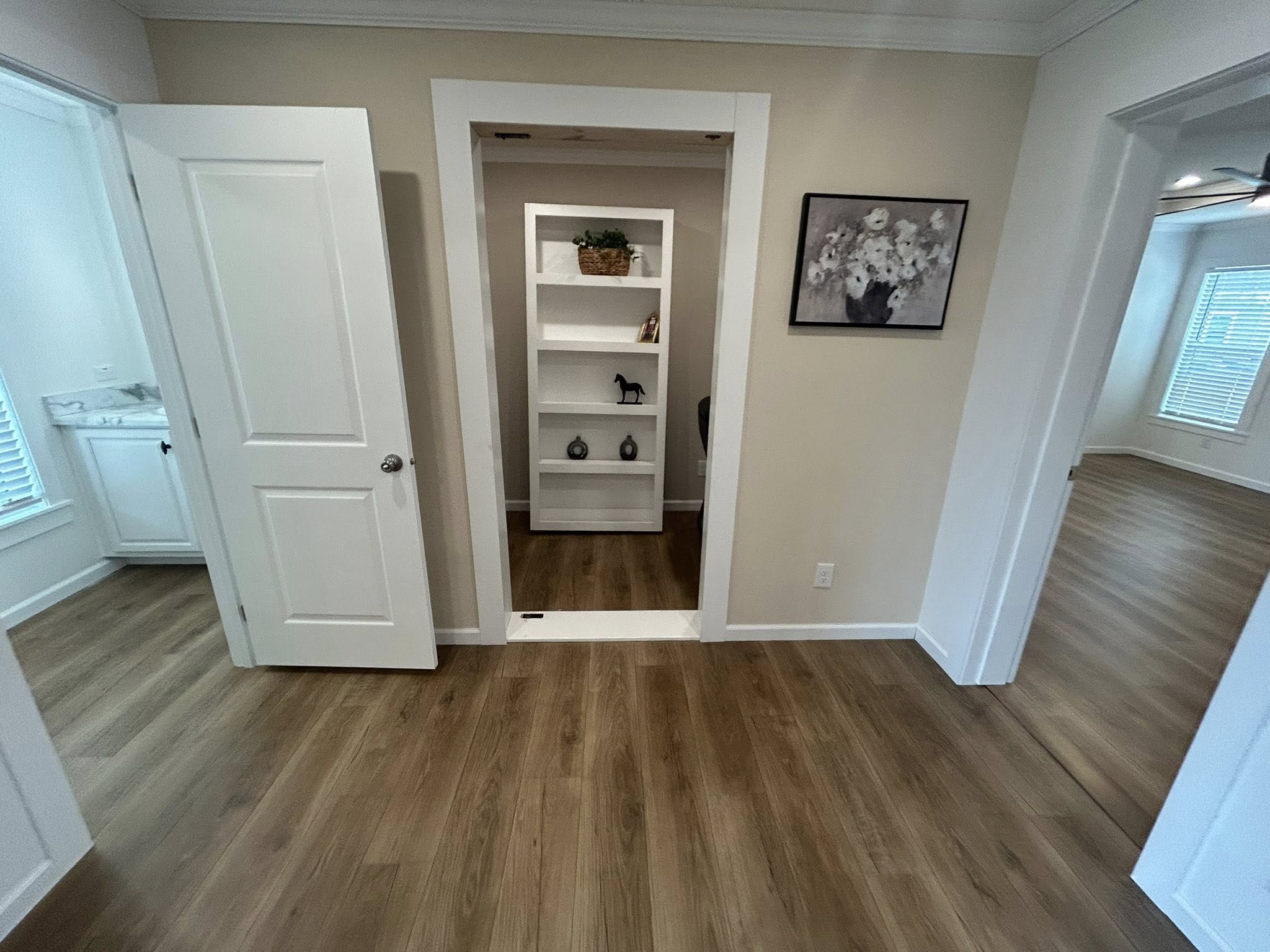 Open room with light wood flooring, white walls, and doorways. A hidden bookshelf door reveals a small room. Art on the wall adds warmth. Cozy atmosphere.