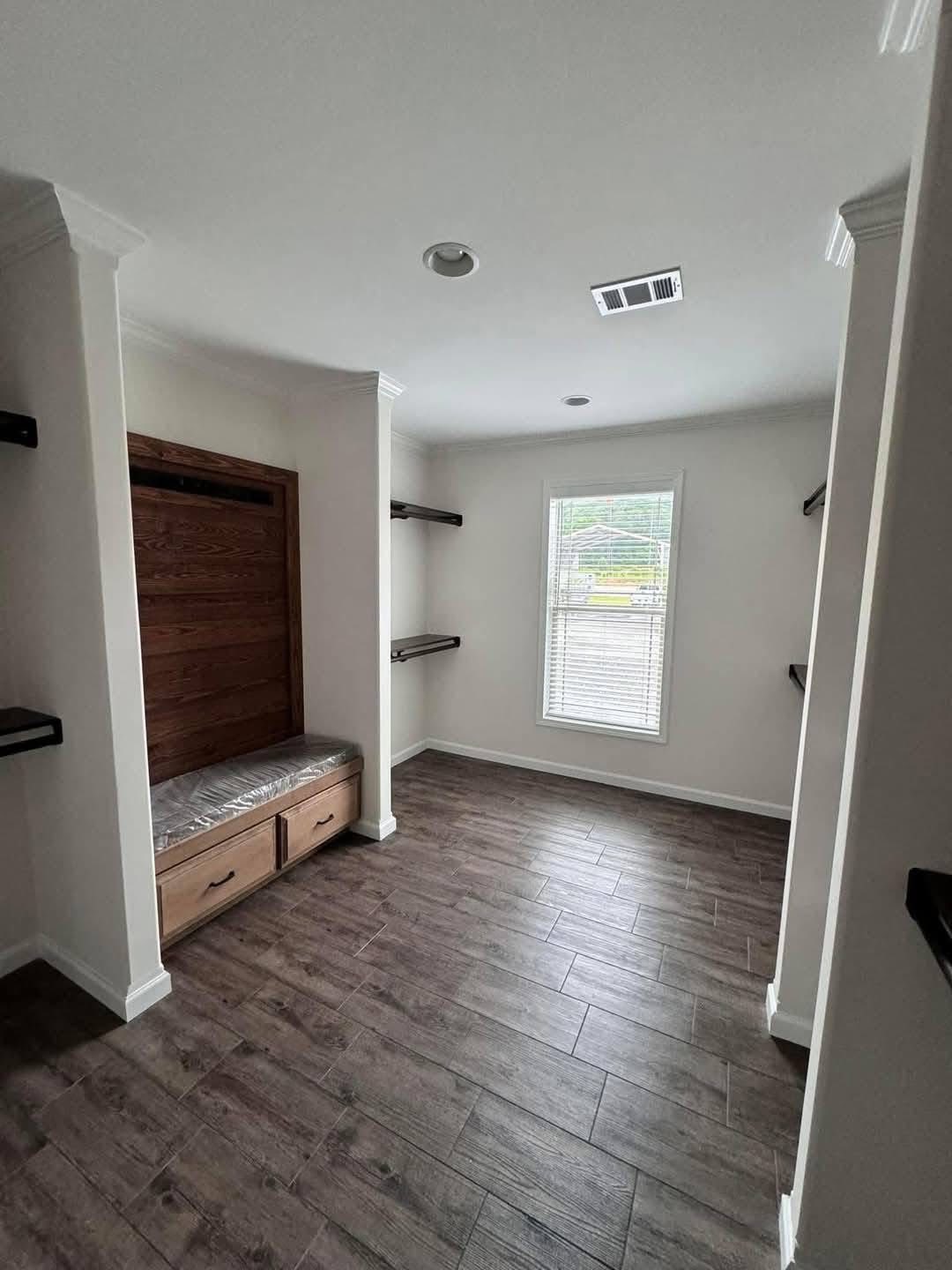 Simple room with wooden flooring, a brown built-in bench with drawers, open shelves, and a window with blinds. It's bright and minimalist.